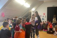 Kinder in Kostümen spielen fröhlich, während Erwachsene im Raum zuschauen und lächeln.